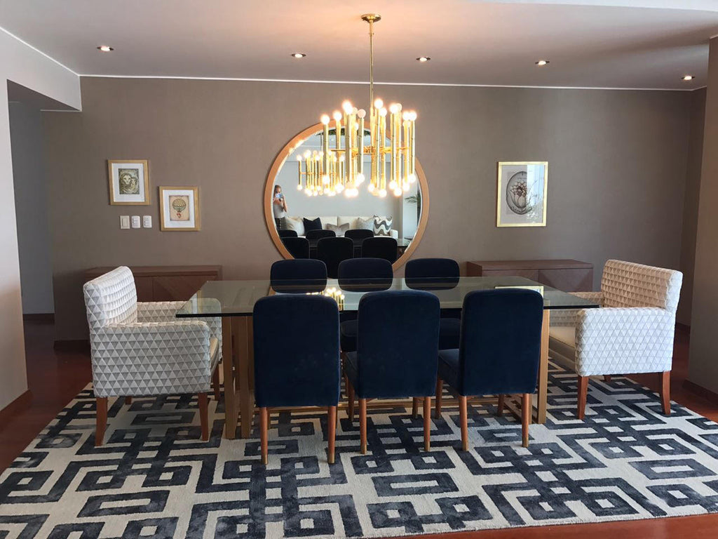 Foto de un comedor con una mesa larga y un candelabro moderno. Dos de las sillas de la mesa son de color blanco y las demás azul oscuro. Detrás de la mesa hay un gran espejo redondo. Debajo de ella hay una alfombre azul y blanca.