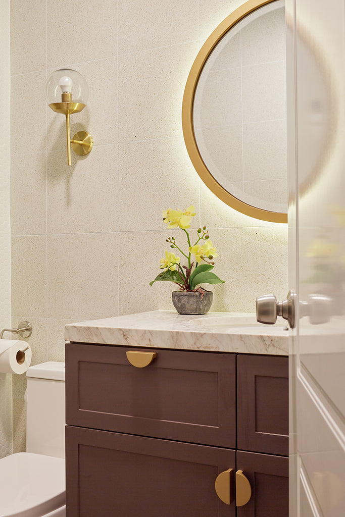 Foto de un baño de visitas. Hay un lavabo, un espejo y un inodoro. Luz sale de atrás del espejo. Sobre el mueble del lavabo hay una flor amarilla.