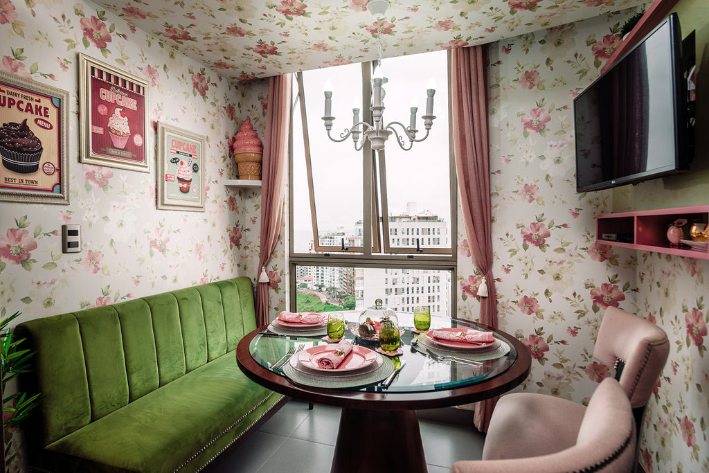Sala de un comedor de diario. Hay una mesa redonda al rededor de la cual hay dos sillas rosadas y un sillón largo verde. Las paredes y el techo están cubiertas de papel decorativo con un patrón floral rosado. Sobre una de las paredes hay una televisión.