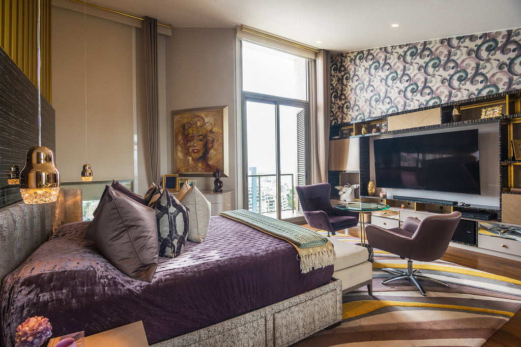 Foto de un dormitorio principal con una cama doble. Hay una pequeña mesa y dos sillas frente a la cama, detrás de las cuales hay un mueble con una televisión. La pared detrás de la televisión tiene papel decorativo en tonalidades moradas.